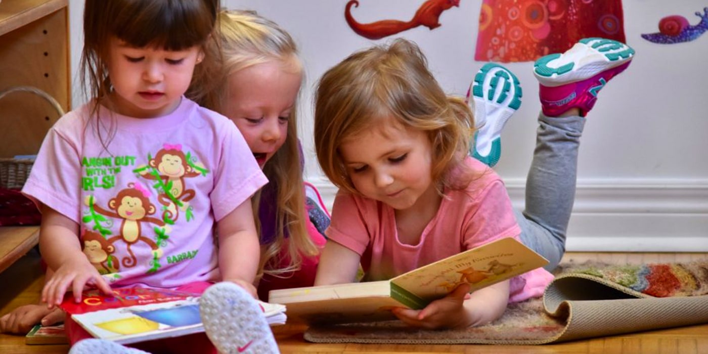 Three toddler age children sitting/lying on the floor looking at books together.