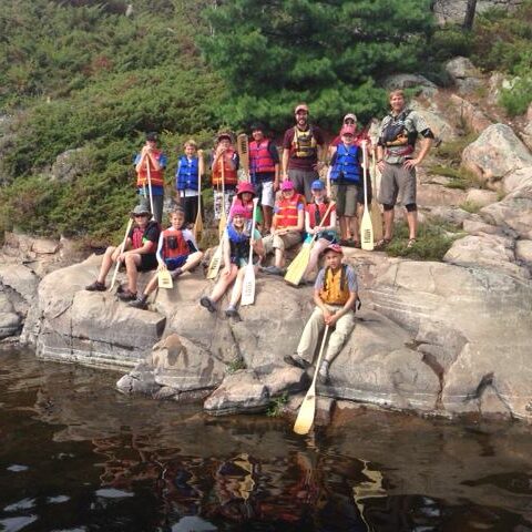 A group of Elementary children sitting on a rock, wearing lifejackets and holding paddles.