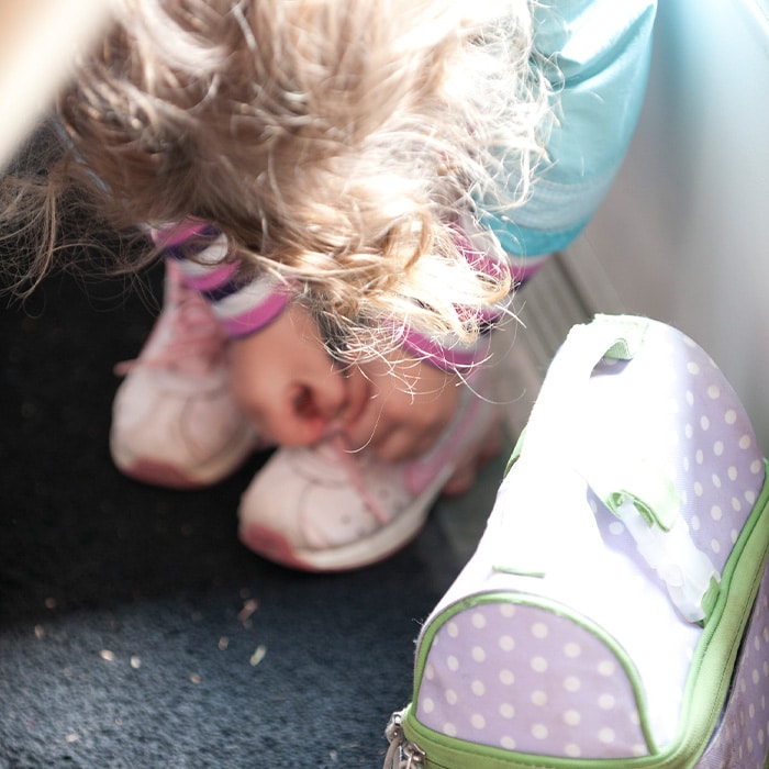 Closeup image of a child's hands as she ties her shoes, with her lunchbox close by.