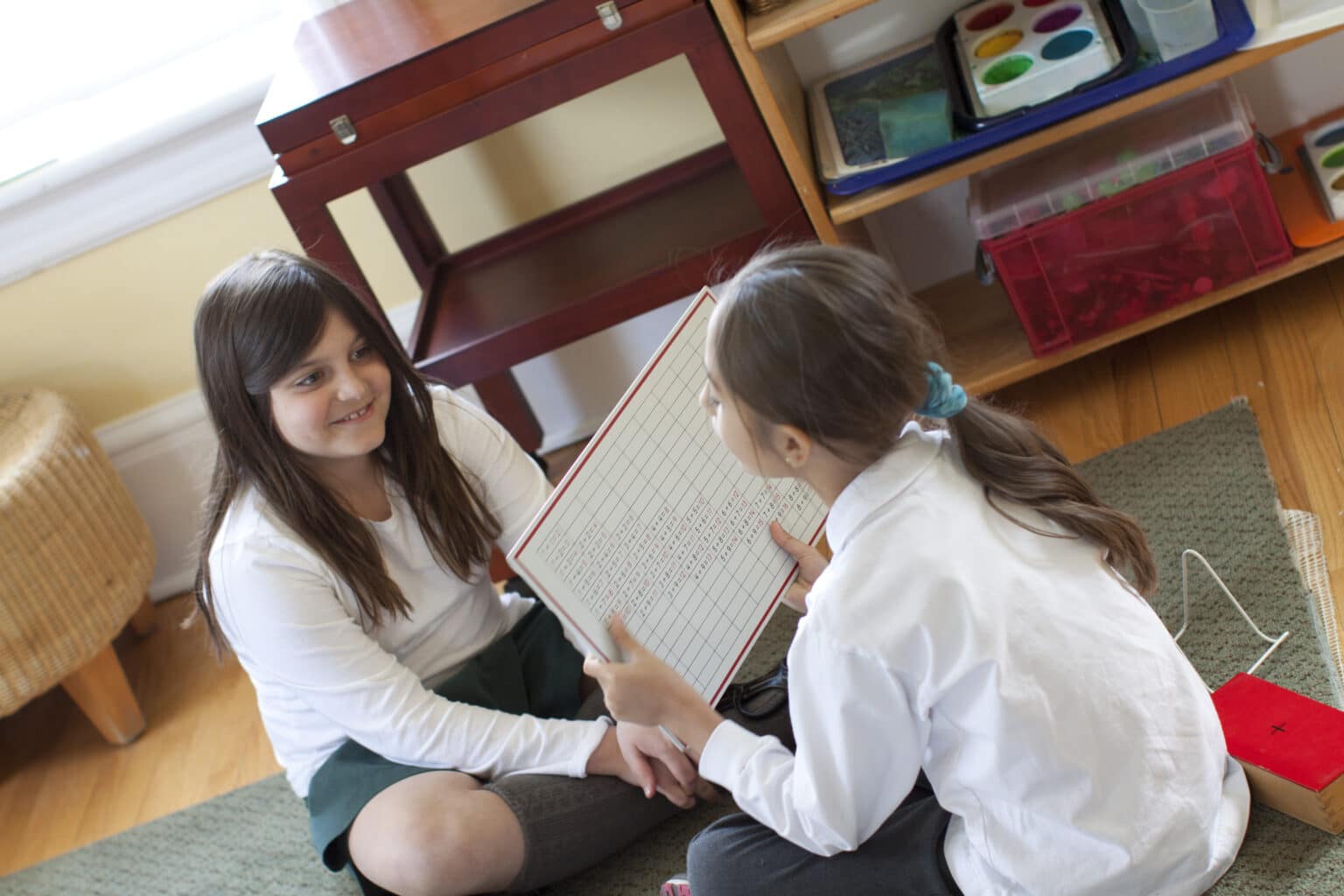 Two lower elementary students working with addition math facts. One is quizzing the other and holding a chart to verify the answers.