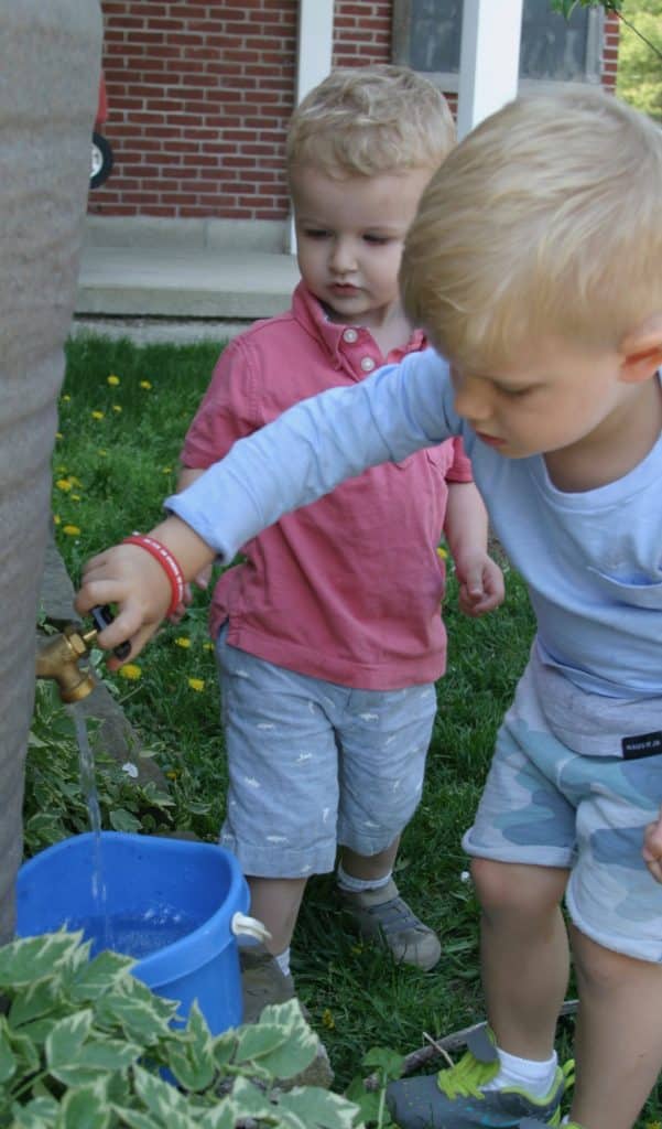 Two toddler students filling a bucket with water from an outdoor faucet.